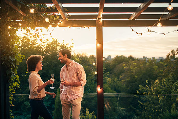 Man and Woman, a couple smiling whilst enjoying a glass of prosecco on a balcony lit by fairy lights, looking over the trees into the city.